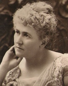 A portrait photograph of a women turned slightly to her right with her right hand touching her face. Her hair is up.
