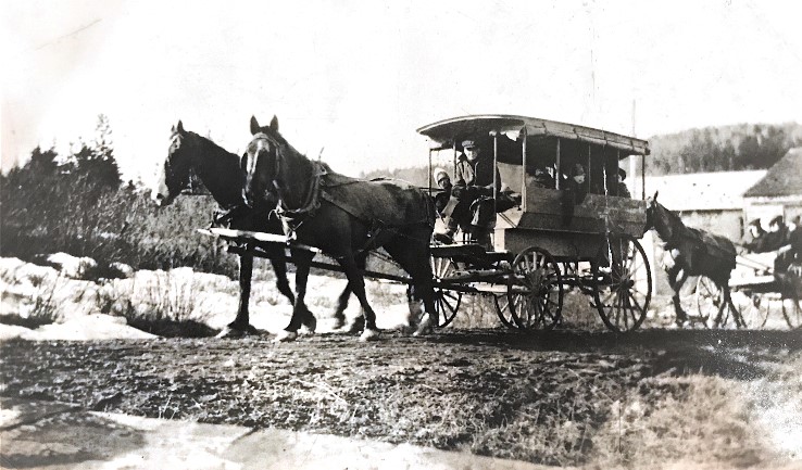 The left view of horse drawn school vans with drivers and children dressed in winter clothing head down a dirt road with snow on either side. The first van is covered and pulled by two horses, the second is uncovered and pulled by one horse.