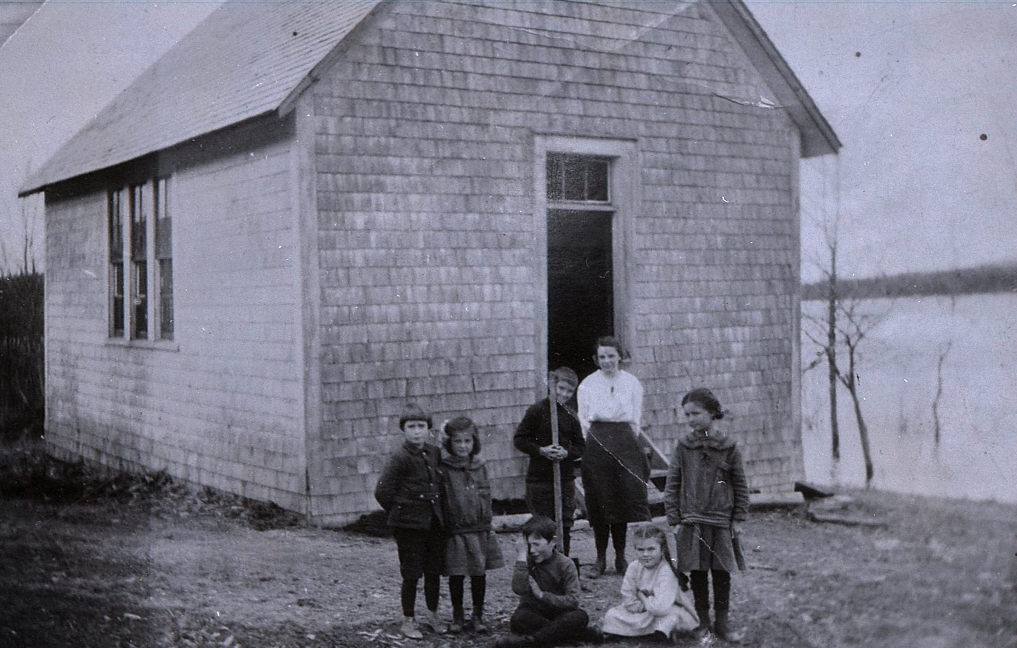 A young teacher and six elementary students in front of a one-room cedar shingle school at the edge of a river in flood,