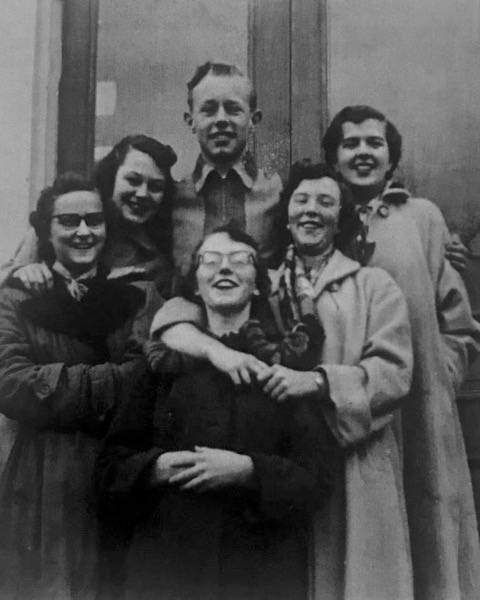 Five young women and one young man wearing light-weight coats smile and pose by a doorway for their high school graduation.