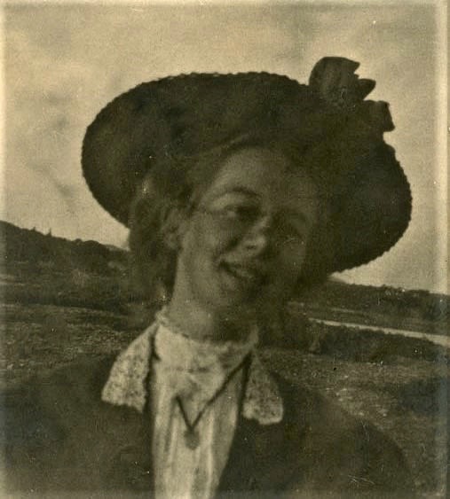 Head and shoulder image of a smiling young woman wearing a high collared blouse, a collared coat and a brimmed hat.