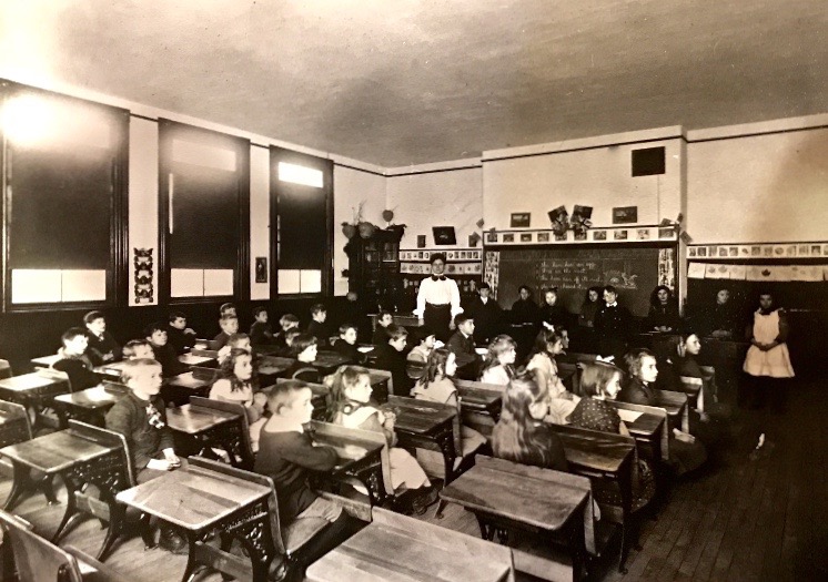 Une salle de classe avec une enseignante et quelques élèves débouts en avant. D’autres jeunes élèves sont assis à leurs pupitres. Sur le mur, il y a des photos de fleurs et de plantes.