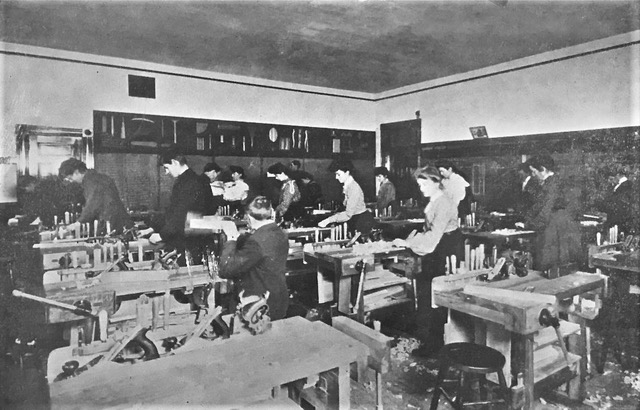 Female students in long skirts and male students in suits at work benches in the manual training room.