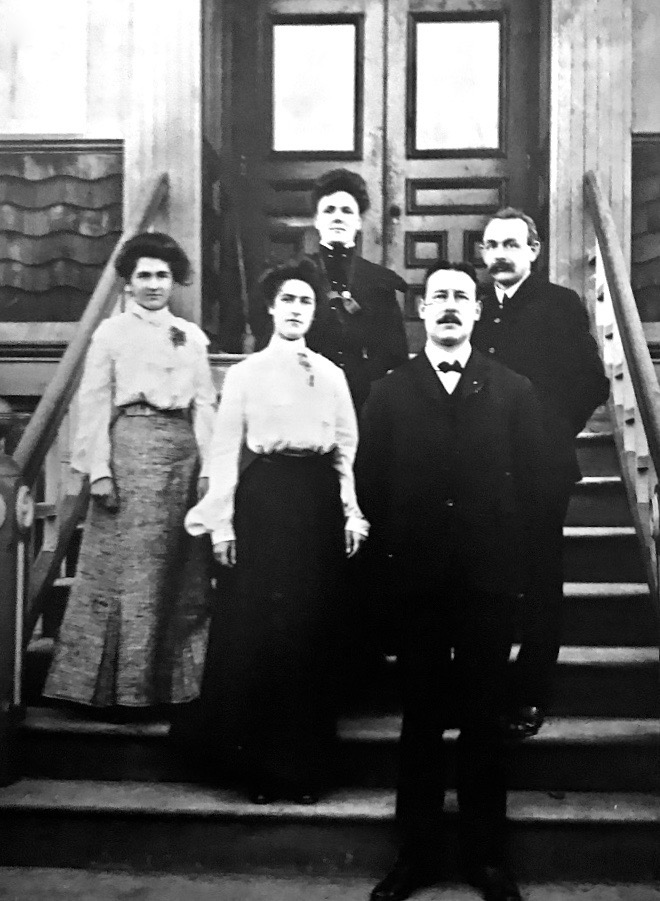 Two female teachers wearing long skirts and two male teaches in suits pose on the front steps of a school.