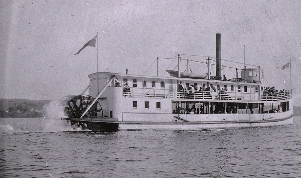 Vue de bâbord d’un bateau à vapeur sur le fleuve. Des passagers se tiennent sur les ponts.