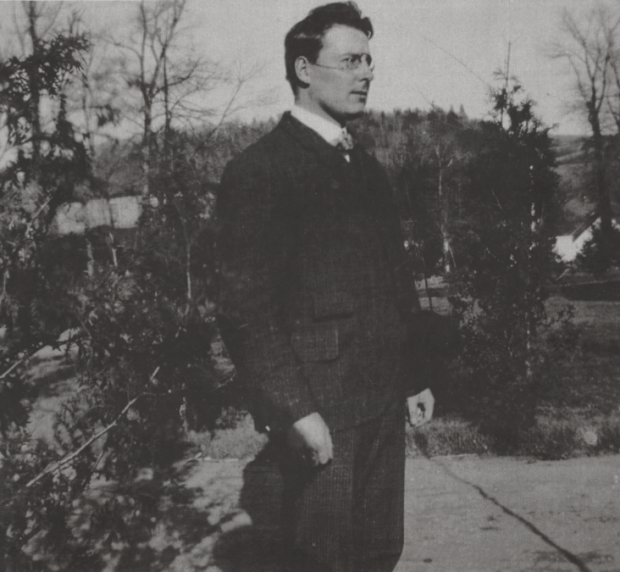 A right side view of teacher as he stands outside the school near cedar trees. He is wearing a suit and glasses.