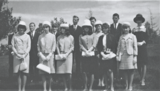 Graduating class includes eight young woman wearing skirt suits and hats. Standing behind them are six young men in suits.