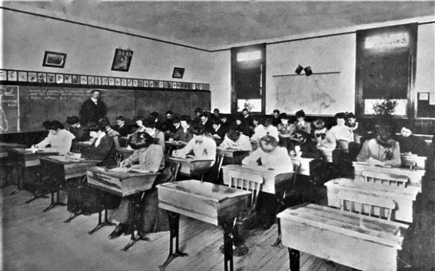 A male teacher with his back to a blackboard has a side view of thirty student who are sitting at their desks completing a test.