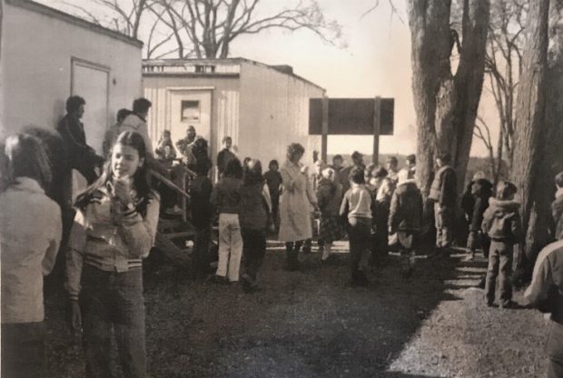 Many students and one teacher take a break on the steps and in front of two portable classrooms.