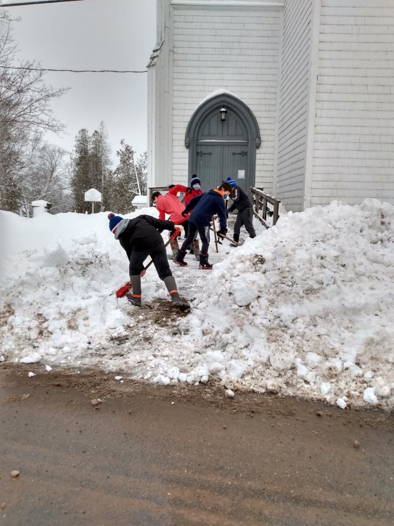 Five students are shovelling deep snow on the walk in front of a church.