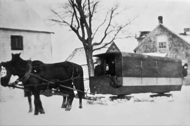 Left side view of a covered school van on runners pulled by two horses, buildings in the background.