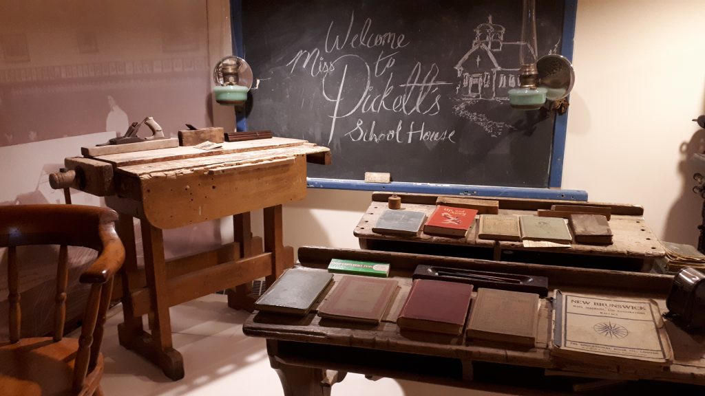 A blackboard, high wooden work bench and two vintage double wooden desks with old school books on them.