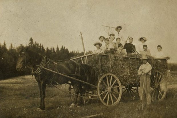 A man with a hay fork stands beside a wagon of hay pulled by a horse. 11 Family members of all ages sit on top of the hay.
