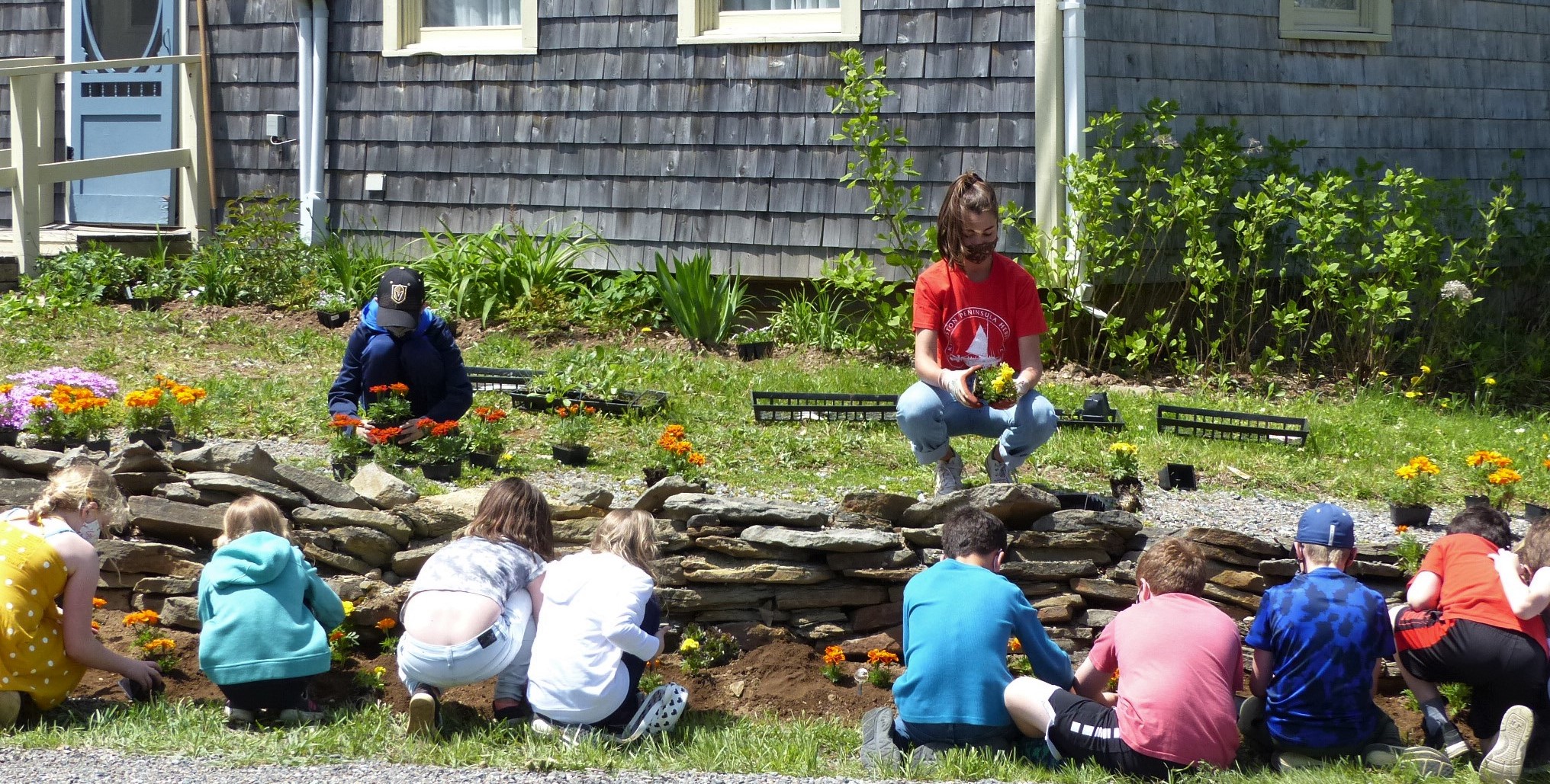 Ten children and a young woman planting flowers in front of a cedar shingled house.