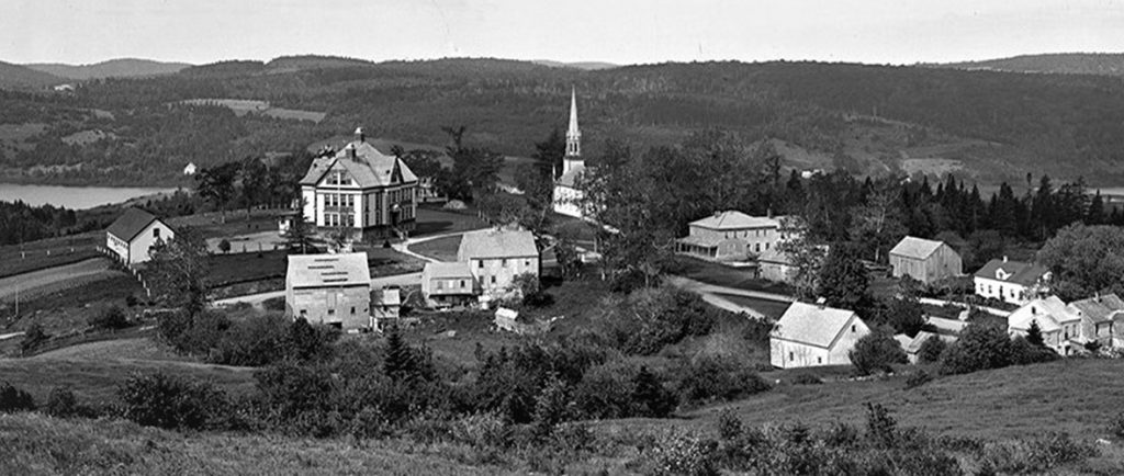 Aerial view of Kingston shows historic MCS , Trinity Church and village buildings set in rolling hills with Bates Lake in the background on the left and Kingston Creek on the right.