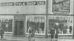Black and white archival photograph. Street view. Window signs from left to right read: CLEARANCE SALE OUR PRICES ARE LOWER. Sign on the right side of the building reads: WINDSOR STYLE SHOP LTD. LEADS IN Fashions WHOLESALE & RETAIL. Six people stand on the sidewalk outside the building. Two boys, two girls, and a mother and daughter.