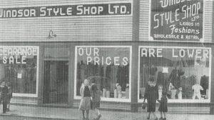 Photographie d'archives en noir et blanc. Vue de la rue. Textes des panneaux aux fenêtres, de gauche à droite : CLEARANCE SALE OUR PRICES ARE LOWER (LIQUIDATION NOS PRIX SONT PLUS BAS). Panneau à droite du bâtiment : WINDSOR STYLE SHOP LTD. LEADS IN Fashions WHOLESALE & RETAIL (MAGASIN WINDSOR STYLE Mode DERNIER CRI VENTE EN GROS ET AU DÉTAIL). Six personnes se tiennent sur le trottoir à l’extérieur du bâtiment. Deux garçons, deux filles et une mère et sa fille.