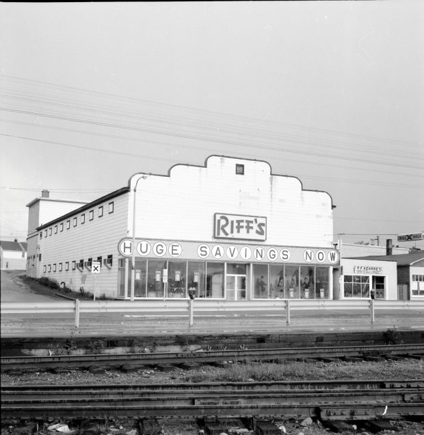 Black and white archival photograph. Street view. Building visible across railway tracks. Sign across building reads RIFF’S and below that HUGE SAVINGS NOW. Male and female manikins are visible in the front windows. P.F. Kearney’s shop is to the right.