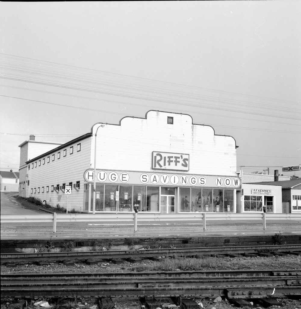 Black and white archival photograph. Street view. Building visible across railway tracks. Sign across building reads RIFF’S and below that HUGE SAVINGS NOW. Male and female manikins are visible in the front windows. P.F. Kearney’s shop is to the right.