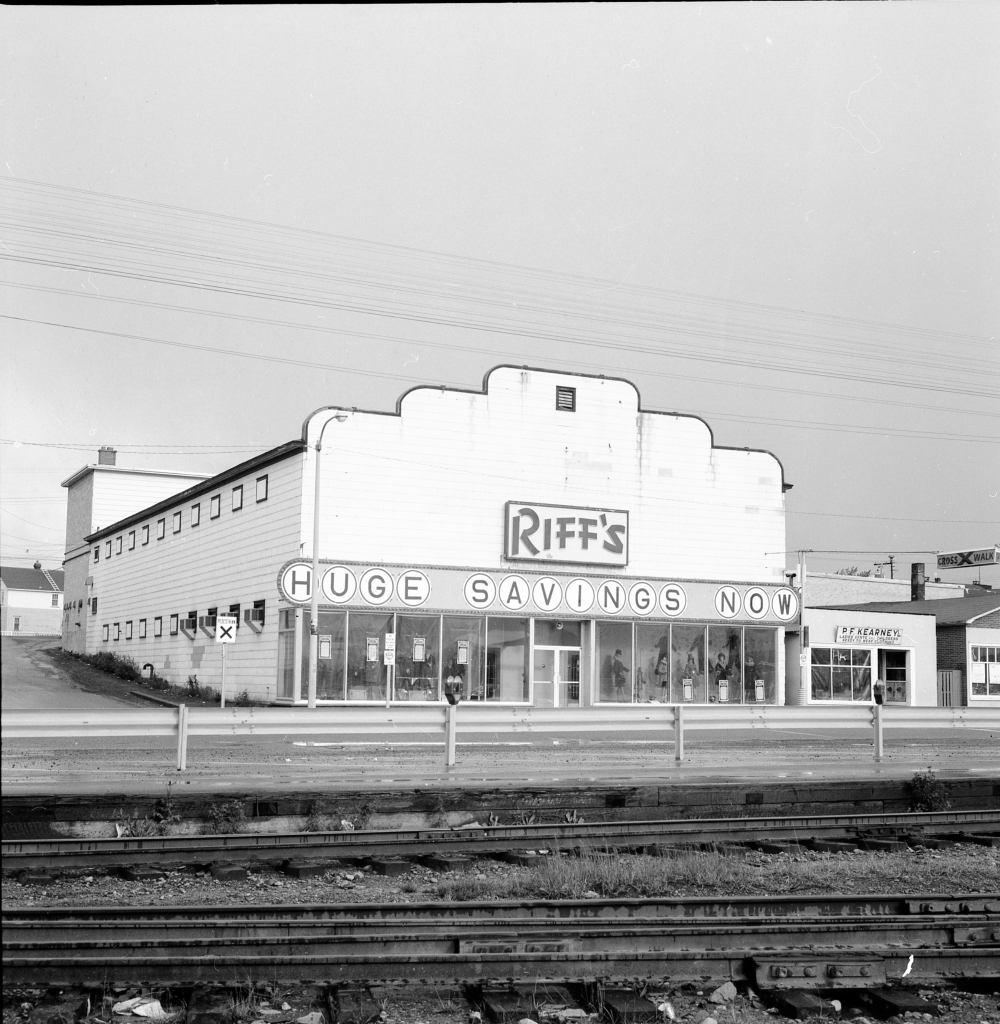 Photographie d'archives en noir et blanc. Vue de la rue. Un bâtiment est visible de l’autre côté des voies de chemin de fer. Texte du panneau accroché en travers du bâtiment : RIFF’S. En dessous : HUGE SAVINGS NOW (GROS RABAIS MAINTENANT). On aperçoit des mannequins hommes et femmes à travers les fenêtres de l’avant du bâtiment. Le magasin P.F. Kearney est à droite.