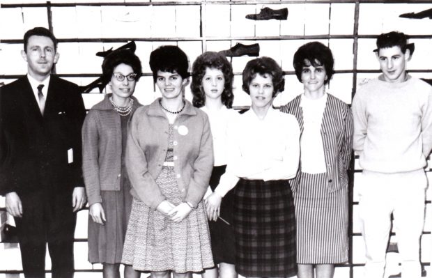 Black and white archival photograph. Staff in front of a wall of shoes. Left to right: man in a business suit with a tie, five women, and a man wearing a long sleeved shirt and pants. All people have dark hair. Women are all wearing skirts and shirts or dresses, and some wear blazers.