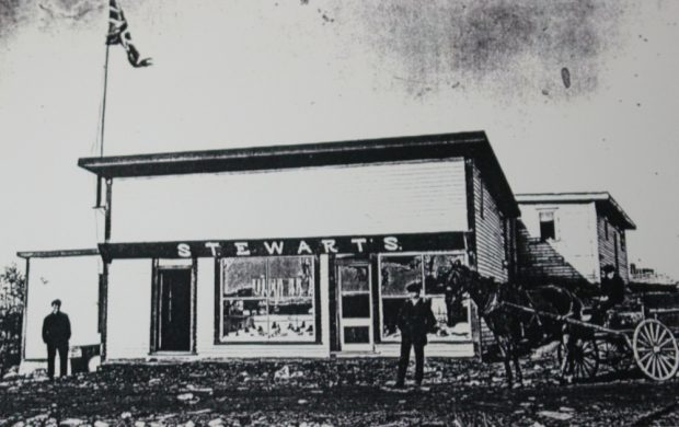 Black and white archival photograph. Street view. The main building has two entrance doors and two large windows. A union jack flies over the left side of building. There are two men standing outside, and another man driving a horse and cart.