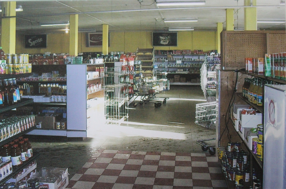 Colour photograph. Several shopping carts line the aisle, cans and bottles line the shelves, and the floor on the front right side is checkered red and white while it is beige in left and back portion of photo.
