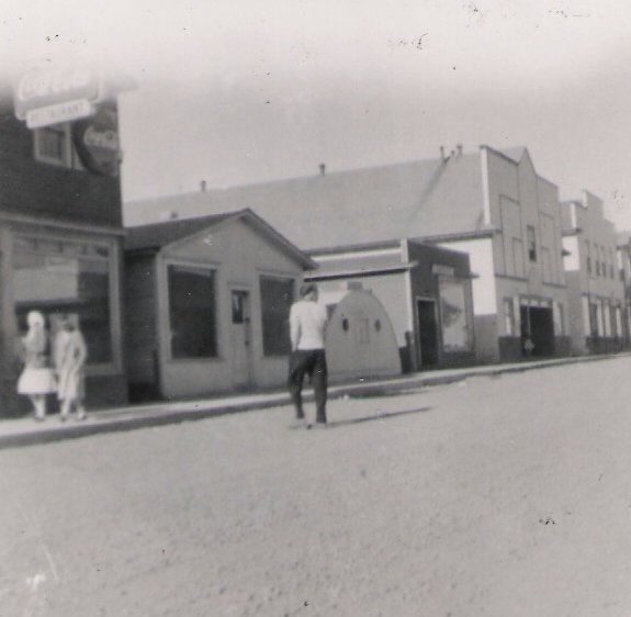 Photographie noir et blanc. Vue de la rue. Vue de la rue Main en direction de l’est. Le bâtiment métallique semi-circulaire (3 e depuis la gauche) est la remorque à frites Stroud’s.