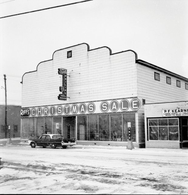 Black and white archival photograph. Street view. Exterior view of Riff’s and P.F. Kearney’s. Christmas décor can be seen in the windows of both shops. Snow is falling and there is a car parked in front of Riff’s. The sign above Riff’s says: RIFF’S CHRISTMAS SALE.
