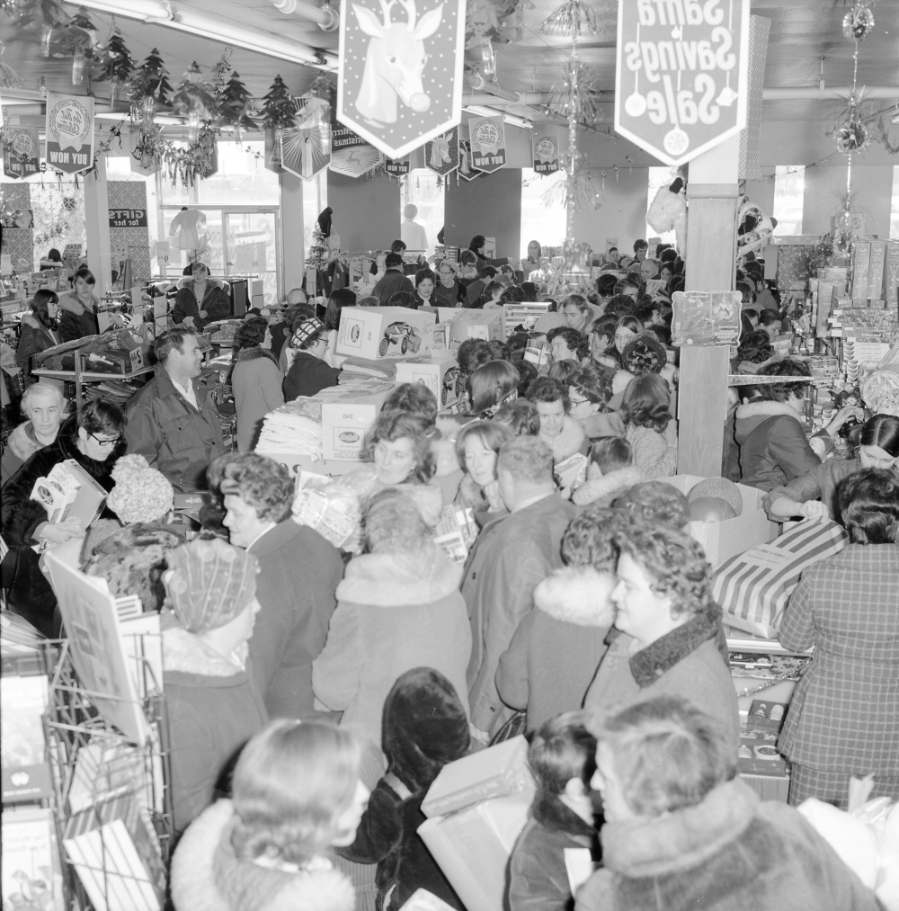 Black and white archival photograph. Over 70 people in winter coats and hats in line at Cohen’s while Christmas decorations and banners hang from the ceiling.