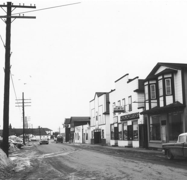 Black and white archival photograph. Street view, Several Main Street businesses on right side. Left side of the street is lined with poles. St. Joseph’s Parish visible in background on left side.