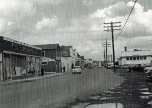 Black and white archival photograph. Street view. Main Street businesses on left side of photo, telephone poles and the railway station on right side. There are four cars on the street, and several pedestrians on the sidewalk in the left part of photo.