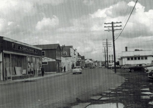 Black and white archival photograph. Street view. Main Street businesses on left side of photo, telephone poles and the railway station on right side. There are four cars on the street, and several pedestrians on the sidewalk in the left part of photo.