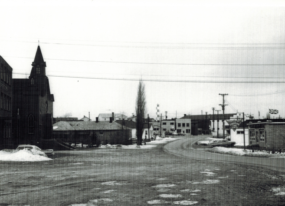 Black and white archival photograph. Street view. Looking north toward Main Street, Windsor from St. Joseph’s Parish which is on the left side of photo. Canadian Tire Corp’n on right side. Railway crossing sign in the centre of the photo.