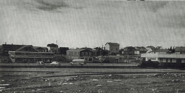 Black and white archival photograph. Street view. Railway tracks in foreground and stores in background. S. Cohen & Sons on left side, train and Newfoundland railway station on right side.