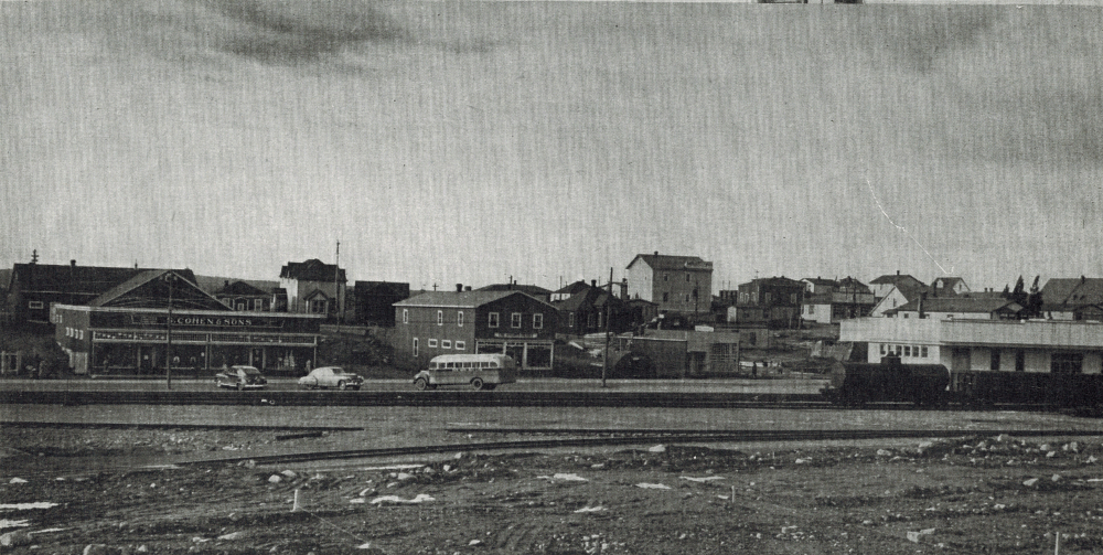 Photographie d'archives en noir et blanc. Vue de la rue. Le chemin de fer au premier plan et magasins en arrière-plan. À gauche on voit le magasin S. Cohen & Fils, à droite le train et la gare du chemin de fer de Terre-Neuve.