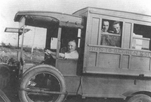Black and white archival photograph. A man drives a bus with two passengers in the first window. The side of the bus reads: BUS TO S. COHEN & SONS.