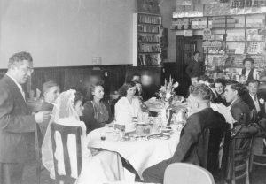 Photographie d'archives en noir et blanc. Participants au mariage de Florence Hollett et Jack Maloney, assis à table dans le Cozy Chat.