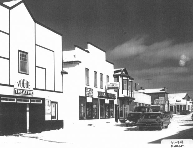 Black and white archival photograph. Street view. Connolly’s Shoe Store and Tuma Jewellers in centre of photo to the right of Vogue Theatre.