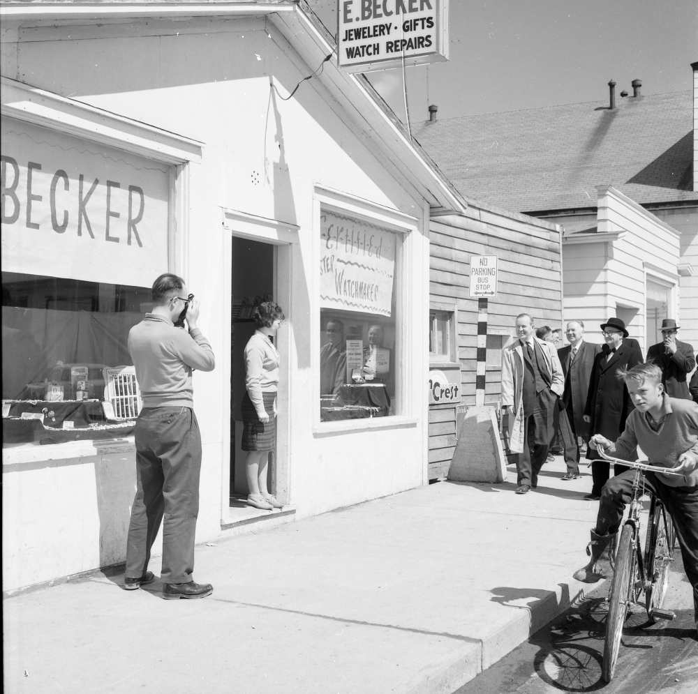 Black and white archival photograph. Street view. Exterior view of E. Becker. Man to the left of photograph taking a picture, woman stands in doorway, group of six men in suits walk towards the shop, and a boy rides his bicycle past shop.