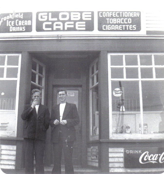 Black and white photograph. Two men standing on steps of the Globe Cafe between two bay windows. Signs on top of building from left to right read: Brookfield ICE CREAM DRINKS, GLOBE CAFE, and CONFECTIONERY, TOBACCO, and CIGARETTES. Bottom right sign says: DRINK Coca-Cola.