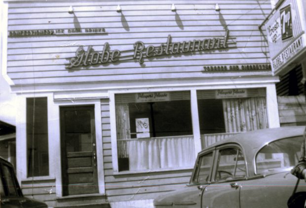 Black and white photograph. Two cars parked in front of the Globe Restaurant. Signs above the door from left to right read CONFECTIONERY ICE COOL DRINKS, Globe Restaurant, and TOBACCO AND CIGARETTES. Sign stating Players Please hung in both windows.