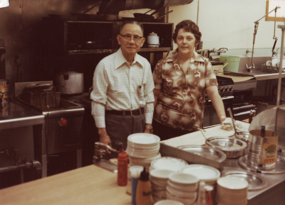 Photographie couleur.  Intérieur de la cuisine du restaurant Globe. On distingue un four derrière l’homme et la femme qui sont au centre de la photo.