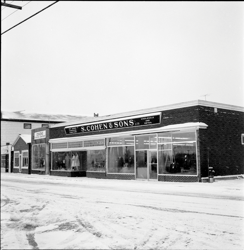 Black and white archival photograph. Street view. S. Cohen & Sons and Cohen’s Furniture Department looking west on Main Street, Windsor. Sign above store reads: DRY GOODS; HARDWARE; FURNITURE; S. COHEN & SONS LTD.; WHOLESALE AND RETAIL. Female and male manikins visible in the store windows.