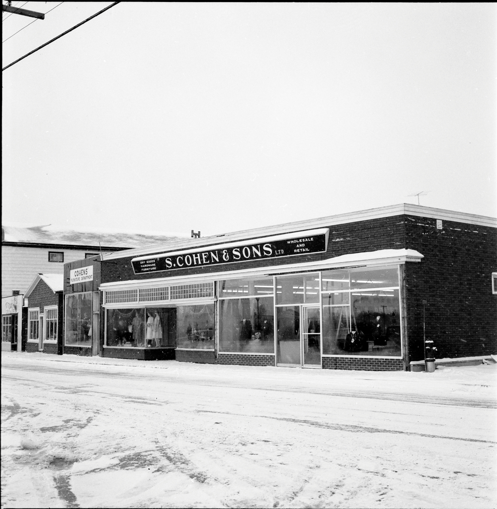 Black and white archival photograph. Street view. S. Cohen & Sons and Cohen’s Furniture Department looking west on Main Street, Windsor. Sign above store reads: DRY GOODS; HARDWARE; FURNITURE; S. COHEN & SONS LTD.; WHOLESALE AND RETAIL. Female and male manikins visible in the store windows.