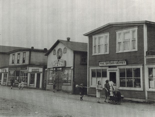 Black and white archival photograph. Street view. Looking west on Main Street, Windsor. Brown Derby, King Edward Theatre, A. Peckford, S. Cohen & Sons, and Purity Cafe visible. Kirk Pomeroy is carrying a birch broom, Daisy Bennett is wearing a light coat, and Mrs. Pomeroy is pushing a stroller.