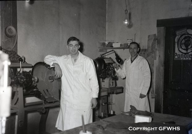 Black and white archival photograph. Two men in white lab coats stand in Hermann Münch’s shoe shop.
