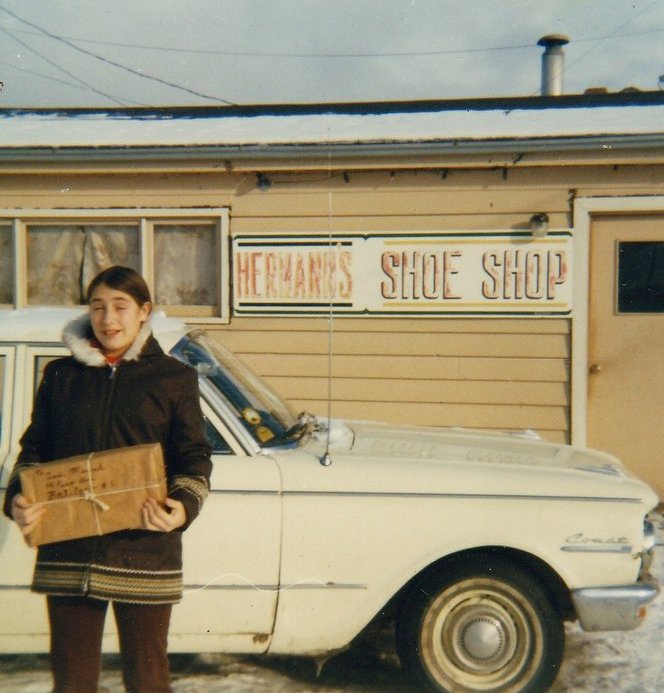 Colour photograph. Street view. Girl in a winter coat holding a package stands in front of a white car outside of Hermann’s Shoe Shop,.