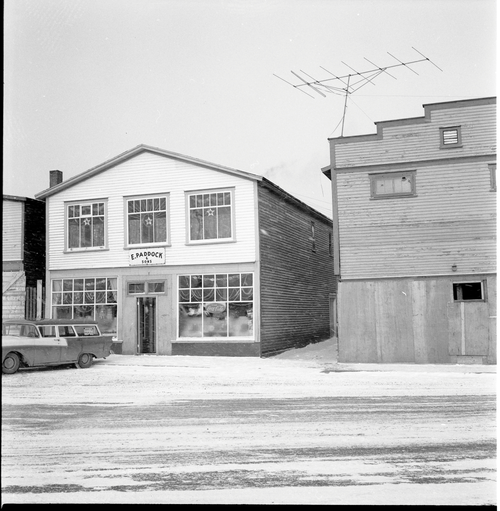 Black and white archival photograph. Street view. Exterior view of E. Paddock & Sons during the Christmas season. Decorative garland and stars can be seen in windows and there are toys displayed in the lower windows.