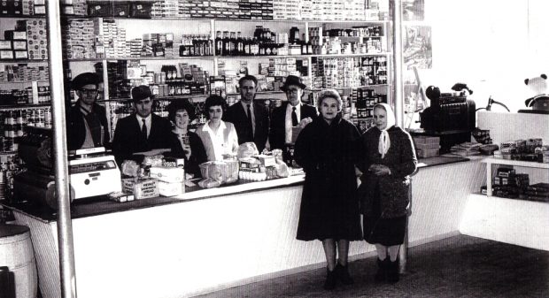 Black and white archival photograph. Four men and two women behind the cash register and counter, and two women in coats in front of the counter. The shelves behind the cash register and stocked full of packages, cans, and bottles of product.